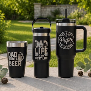 May include: Three matte black stainless steel drink containers. The left tumbler reads DAD NEEDS A BEER with a beer mug icon. The center bottle reads DAD LIFE IS THE BEST LIFE. The right tumbler features a handle, straw, and THE BEST PAPA IN THE WORLD.