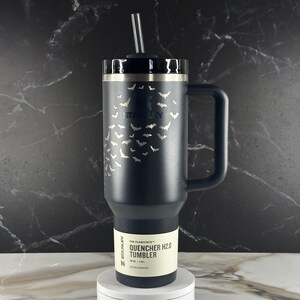 May include: A matte black Stanley Quencher H2.0 tumbler with a 40 oz capacity, featuring a laser-etched pattern of small flying bats across the upper body. The insulated cup includes a sturdy side handle, a black lid, and a clear plastic straw.