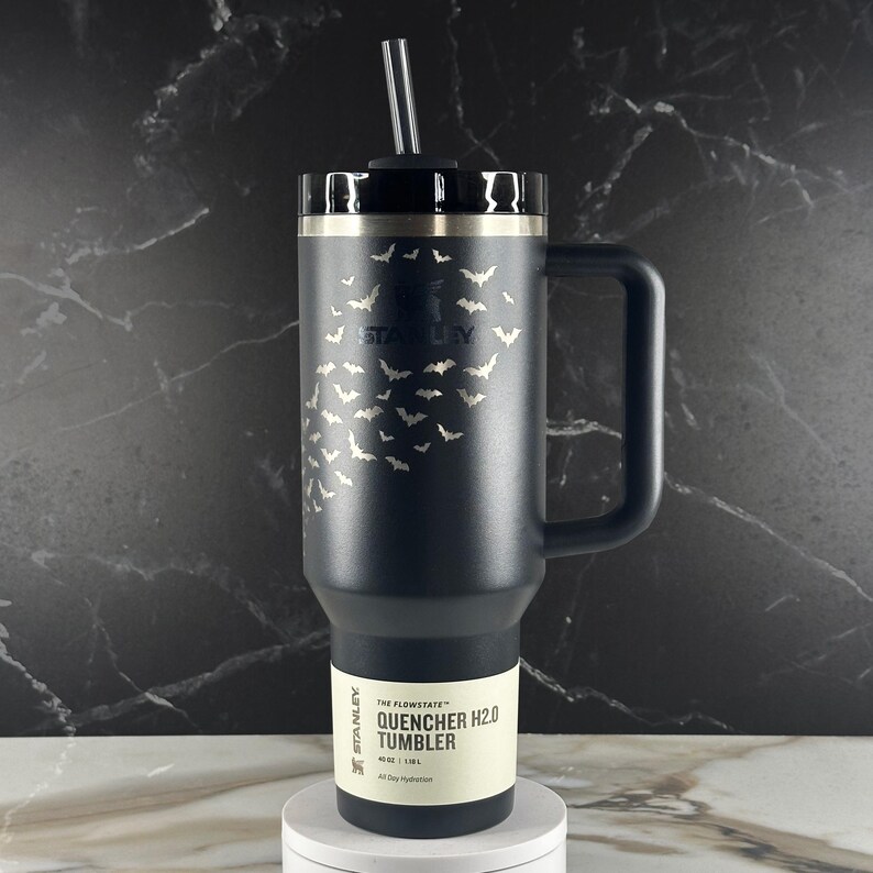 May include: A matte black Stanley Quencher H2.0 tumbler with a 40 oz capacity, featuring a laser-etched pattern of small flying bats across the upper body. The insulated cup includes a sturdy side handle, a black lid, and a clear plastic straw.