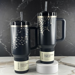 May include: Two matte black Stanley Quencher H2.0 tumblers with handles, lids, and clear straws. The left tumbler features a pattern of small bats, while the right tumbler displays a spiderweb design. Both have labels reading The Flowstate Quencher H2.0.