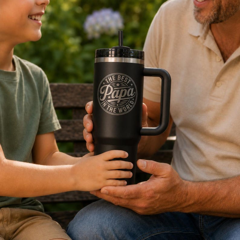 May include: A matte black stainless steel tumbler with a handle, a lid, and a straw. The front features a circular engraved design with the text "THE BEST PAPA IN THE WORLD" surrounding the word "Papa" in a stylized script font.