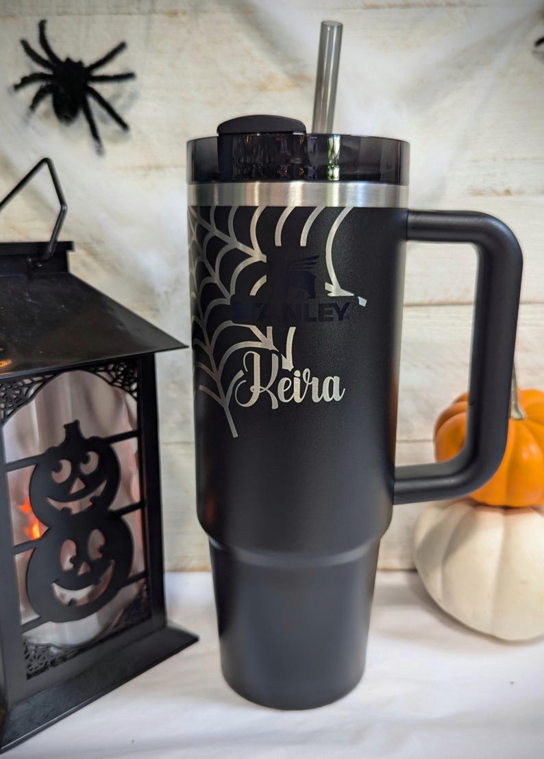 A black 30oz Stanley Quencher Tumbler engraved with a beautiful spider web across the top of the cup with the name Keira across it. The spider web engraving and name dynamically meshes with the Stanley Logo embedded in the tumbler.
