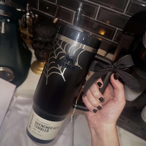 Woman with black nails holding a black 30oz Stanley Quencher Tumbler engraved with a beautiful spider web across the top of the cup. The spider web dynamically meshes with the Stanley Logo embedded in the tumbler.