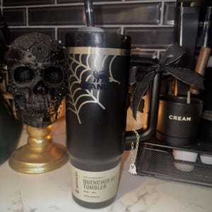A black 30oz Stanley Quencher Tumbler engraved with a beautiful spider web across the top of the cup placed within a Halloween setting. The spider web engraving dynamically meshes with the Stanley Logo embedded in the tumbler.