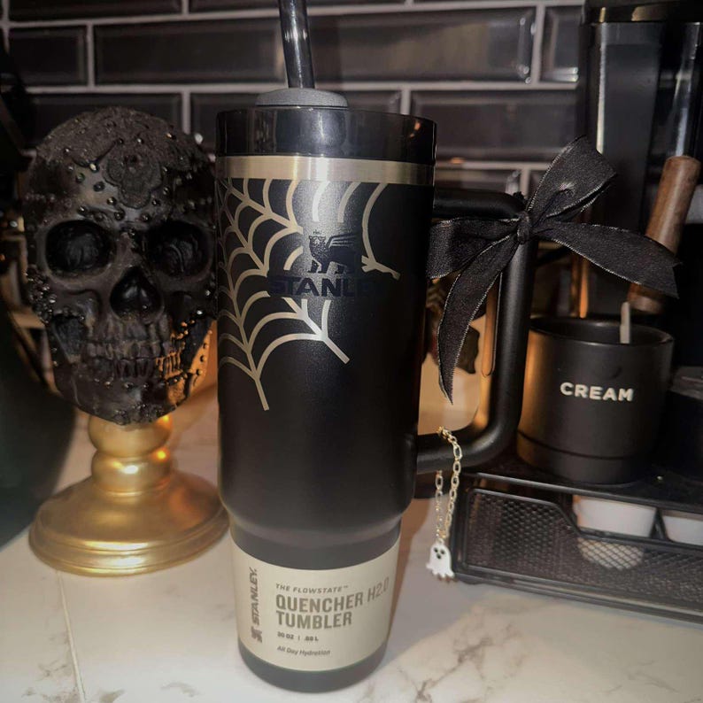 A black 30oz Stanley Quencher Tumbler engraved with a beautiful spider web across the top of the cup placed within a Halloween setting. The spider web engraving dynamically meshes with the Stanley Logo embedded in the tumbler.