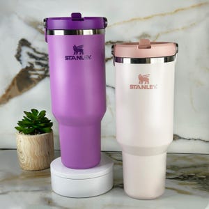May include: Two Stanley brand insulated tumblers. One is a vibrant purple with a matching lid, the other is a soft pink with a light pink lid. Both have a silver rim and the Stanley logo.