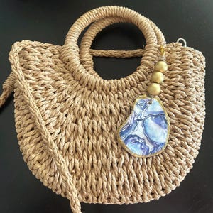 May include: A woven straw handbag with a round top handle and a detachable shoulder strap. The bag features a decorative charm with a blue and white abstract design and gold-colored beads. The bag is tan in color.