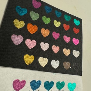 Puede incluir: Una tarjeta negra con 25 muestras de pintura en forma de coraz&oacute;n en varios colores met&aacute;licos. La tarjeta est&aacute; etiquetada con los nombres de los colores, incluyendo Chrome Purple, Chrome Blue, Chrome Light Blue, Chrome Emerald y Chrome Teal.