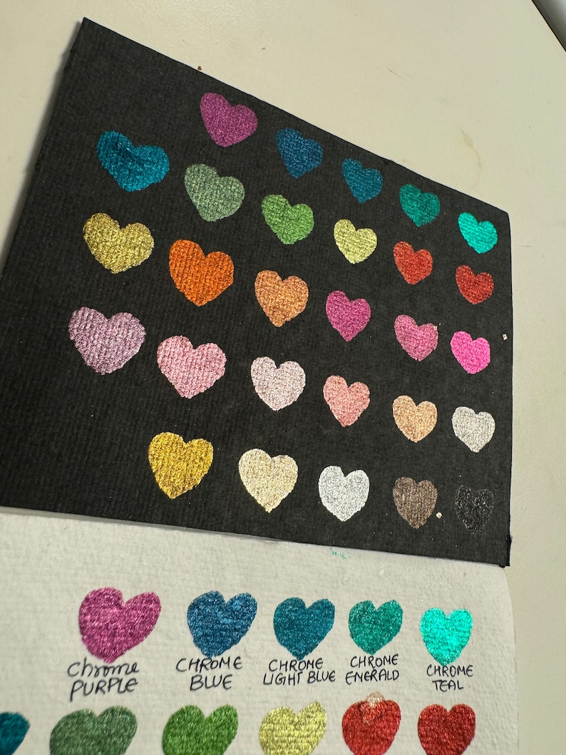 Puede incluir: Una tarjeta negra con 25 muestras de pintura en forma de coraz&oacute;n en varios colores met&aacute;licos. La tarjeta est&aacute; etiquetada con los nombres de los colores, incluyendo Chrome Purple, Chrome Blue, Chrome Light Blue, Chrome Emerald y Chrome Teal.