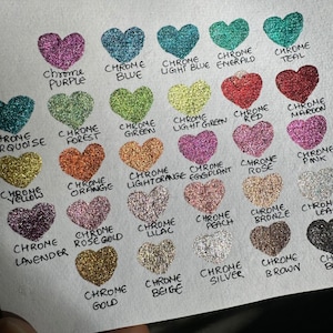 Puede incluir: Una colecci&oacute;n de 20 peque&ntilde;os contenedores en forma de coraz&oacute;n llenos de purpurina de diferentes colores. Cada contenedor est&aacute; etiquetado con el nombre del color de la purpurina, como "Chrome Purple", "Chrome Blue", "Chrome Teal" y "Chrome Gold".