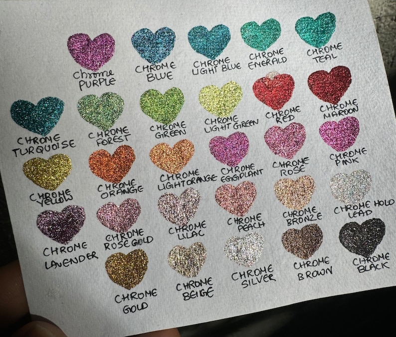 Puede incluir: Una colecci&oacute;n de 20 peque&ntilde;os contenedores en forma de coraz&oacute;n llenos de purpurina de diferentes colores. Cada contenedor est&aacute; etiquetado con el nombre del color de la purpurina, como "Chrome Purple", "Chrome Blue", "Chrome Teal" y "Chrome Gold".