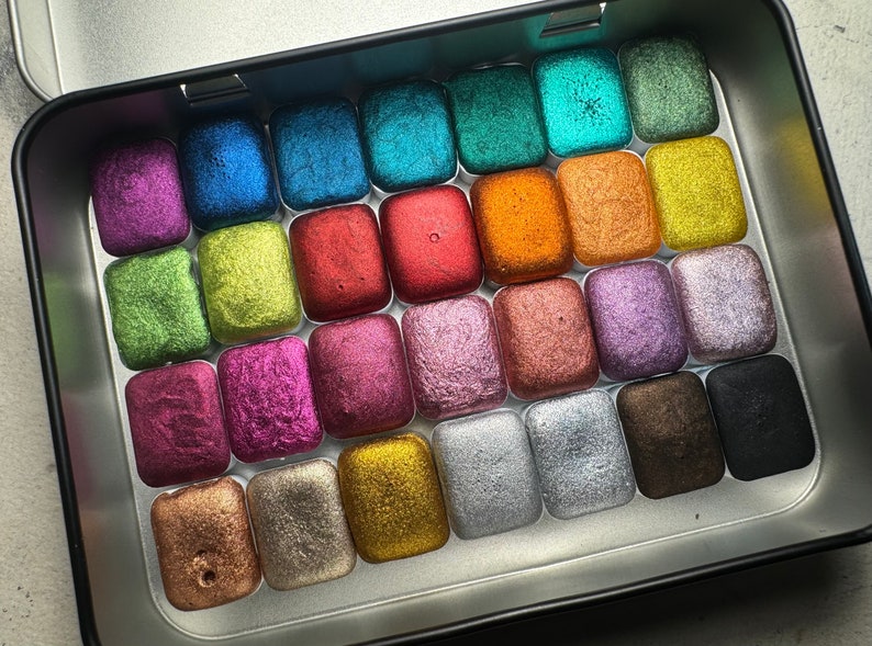 Puede incluir: Una lata de metal llena de 24 peque&ntilde;os cuadrados de pintura acuarela met&aacute;lica. La pintura est&aacute; dispuesta en filas y columnas, y cada cuadrado tiene un color diferente. Los colores incluyen oro, plata, cobre, bronce, verde, azul, morado, rosa, rojo y negro.