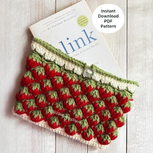 May include: A handmade crochet book sleeve featuring a pattern of red strawberries with green leaves on a white background. The top edge is trimmed with green and white crochet. A book titled "Blink" is partially visible.