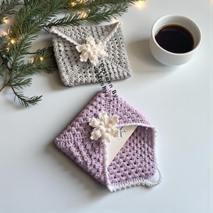 May include: Two crocheted square envelopes, one gray and one lavender, each adorned with a white snowflake. The lavender envelope holds a card. A cup of coffee and a sprig of evergreen are in the background.