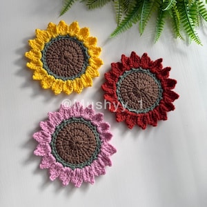 May include: Three crocheted sunflower coasters in yellow, red, and pink. Each coaster has a brown centre, a green ring, and petals in the respective colours. The coasters are handmade and decorative.