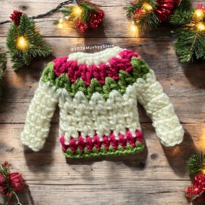 May include: A white crocheted sweater with green and pink stripes. The sweater has long sleeves and a crew neck. The sweater is on a wooden background with Christmas lights and greenery.