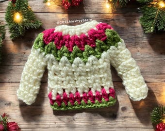 May include: A white crocheted sweater with green and pink stripes. The sweater has long sleeves and a crew neck. The sweater is on a wooden background with Christmas lights and greenery.