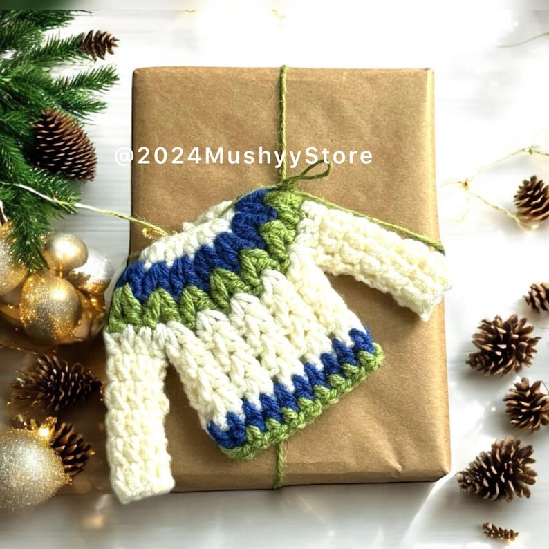 Pu&ograve; includere: Un ornamento a forma di maglione bianco lavorato a uncinetto con strisce verdi e blu. L'ornamento &egrave; appeso a una scatola regalo avvolta in carta marrone con spago.