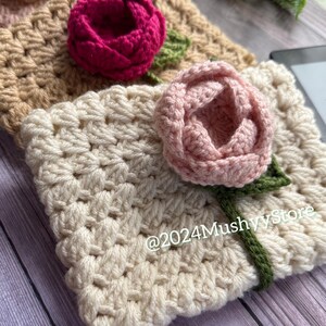 May include: Three crocheted flower embellishments in beige, pink, and red. The flowers are attached to a beige crocheted fabric with green stems.