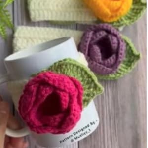 May include: A white mug with a pink crochet rose and green leaf, with a cream-colored crochet band. Additional crochet roses in yellow and purple with green leaves are also visible. The text "Pattern Designed By - @Mushyy.1" is on the mug.