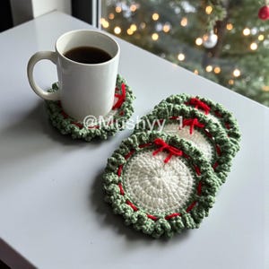 Peut inclure: Une tasse blanche contenant une boisson sombre repose sur un dessous de verre crocheté. Le dessous de verre a un centre blanc, une bordure verte et des détails rouges. D'autres dessous de verre sont empilés, chacun avec un nœud rouge.