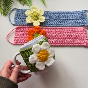 May include: Crocheted daisy-themed items, including face masks in blue and pink, and a green mug cozy. The daisy flowers have white petals and yellow centers. The mug cozy is wrapped around a white mug.