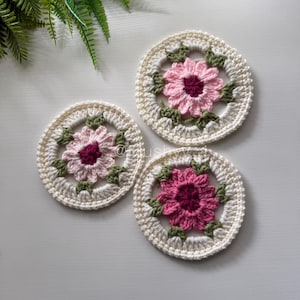 May include: Three handmade crochet coasters. Each coaster features a central pink flower with a dark centre, surrounded by green leaves and a cream-coloured border. The coasters are arranged on a white surface, showcasing the intricate floral design.
