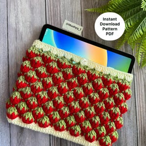 May include: A crocheted tablet sleeve featuring a repeating pattern of red strawberries with green leaves. The sleeve is cream-colored with a green border and holds a tablet. Text on the image reads "Instant Download Pattern PDF".