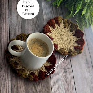 Può includere: Due sottobicchieri all'uncinetto in tonalità di marrone, verde e rosso. I sottobicchieri sono rotondi e hanno un design floreale. Una tazza da caffè bianca si trova sopra uno dei sottobicchieri.