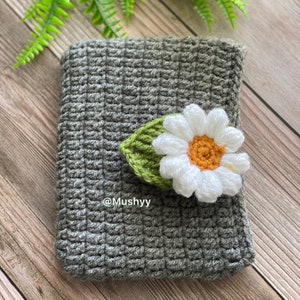 May include: A gray crocheted blanket with a white crocheted daisy flower and green leaf on top. The text "@Mushyy" is visible on the blanket.