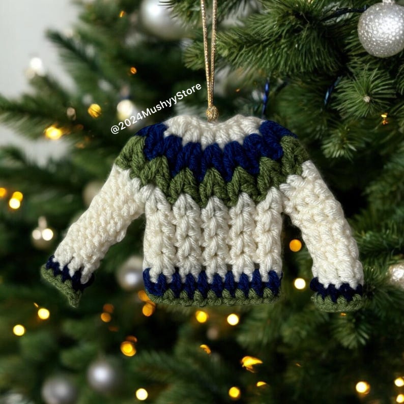 Pu&ograve; includere: Un ornamento a forma di maglione lavorato a uncinetto in bianco, verde e blu appeso a un albero di Natale. L'ornamento ha la forma di un maglione con sfondo bianco e strisce verdi e blu.