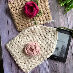 May include: A crocheted Kindle cover with a pink flower and a green strap. The cover is made with a white yarn and has a square stitch pattern. The cover is lying on a wooden surface with a Kindle e-reader underneath. The text on the Kindle screen reads "books on deal".