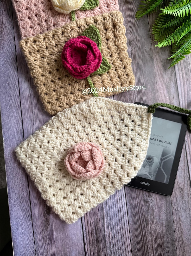May include: A crocheted Kindle cover with a pink flower and a green strap. The cover is made with a white yarn and has a square stitch pattern. The cover is lying on a wooden surface with a Kindle e-reader underneath. The text on the Kindle screen reads "books on deal".