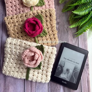 May include: Three crocheted book covers with flower embellishments in pink, beige, and white. The covers are stacked on a wooden surface with a black Kindle e-reader displaying the text "Top eBooks on deal Kindle Monthly Book Deals".
