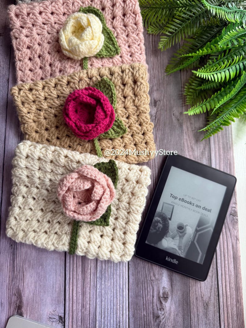 May include: Three crocheted book covers with flower embellishments in pink, beige, and white. The covers are stacked on a wooden surface with a black Kindle e-reader displaying the text "Top eBooks on deal Kindle Monthly Book Deals".