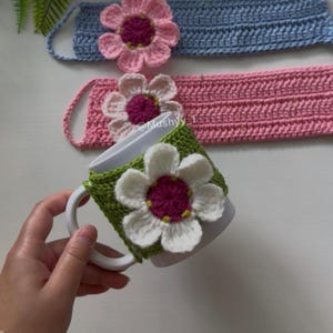 以下が含まれることがあります： 大きな白とマゼンタの花が付いた緑色のかぎ針編みのカバーが付いた白いセラミックマグ。かぎ針編みの花が付いた2つのヘッドバンド（ピンクとブルー）が背景にあります。手作りのアイテムです。