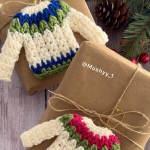 Pu&ograve; includere: Due ornamenti a forma di maglione all'uncinetto, uno con strisce verdi e blu e l'altro con strisce verdi e rosa, sono esposti su carta da regalo marrone. Il testo "@Mushyy_1" &egrave; visibile sulla carta da regalo.
