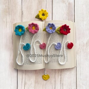 May include: A white crocheted bookmark with colourful crocheted flowers and hearts. The bookmark is attached to a book with a white cover.