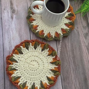 May include: Two crocheted coasters with a white centre and a multi-coloured border in shades of orange, green, and brown. The coasters are on a wooden surface. A white mug with coffee is sitting on one of the coasters.
