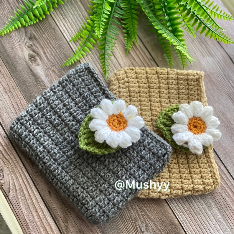 May include: Two crocheted headbands with a daisy flower embellishment. One headband is gray and the other is beige. The headbands are on a wooden surface with green ferns.
