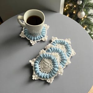May include: A white mug filled with dark liquid sits on a blue and white crocheted coaster. Several similar coasters are stacked nearby. The coasters have a circular blue center with a white scalloped edge.