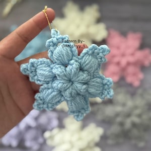 Peut inclure: Ornement en forme de flocon de neige crocheté bleu clair, avec un fil doré pour suspendre. Le flocon de neige est fait de fil doux et présente un design texturé en trois dimensions. D'autres flocons de neige de différentes couleurs sont visibles en arrière-plan. Le texte sur l'image indique "Pattern By - @Mushyy_1".