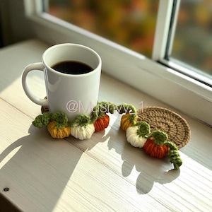 Puede incluir: Una taza blanca llena de líquido oscuro descansa sobre un posavasos de calabaza de ganchillo. El posavasos presenta una cadena de calabazas de ganchillo en naranja, amarillo y blanco con hojas verdes. También hay un segundo posavasos redondo marrón.