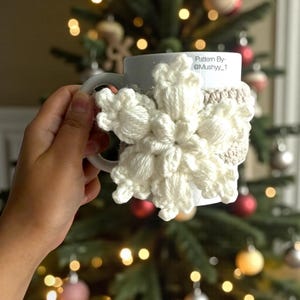 May include: A white ceramic mug adorned with a large, cream-colored, crocheted snowflake and a beige cozy. The mug is held in front of a Christmas tree with ornaments and lights. Text on the mug reads "Pattern By- @Mushyy_1".
