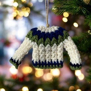 May include: A handcrafted Christmas tree decoration in the shape of a small sweater. The crochet design features an off-white base with blue and green chevron patterns. It has a gold hanging string.