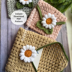 May include: Three crocheted envelope pouches in grey, pink, and tan, each decorated with a white daisy with an orange centre and green leaves. The tan pouch is open, revealing a card. The image includes the text "Instant Download PDF Pattern".
