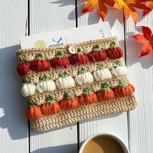 Pode incluir: Uma capa de livro de crochê bege adornada com fileiras de abóboras 3D vermelhas, brancas e laranja, com caules verdes. Um botão de madeira prende a aba. A capa contém um livro, com folhas de outono ao fundo.