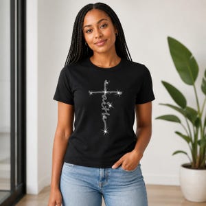 May include: A black short-sleeved t-shirt featuring a silver glittery cross graphic centered on the chest. The word faith is written vertically in a cursive script within the cross design, which includes small star-like accents.