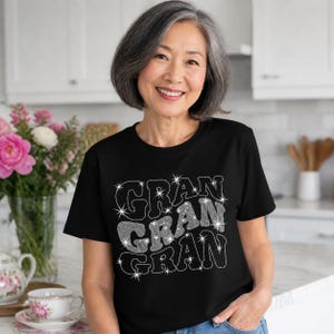 以下が含まれることがあります： 黒の半袖Tシャツで、前面に「GRAN」という単語が3回重ねて配置されたスタイリッシュな白いフォントでプリントされています。文字にはキラキラとした質感があり、デザイン全体に小さな白い星のアクセントが散りばめられています。