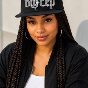 Puede incluir: Una gorra de béisbol negra con el texto "big rep" en pedrería plateada brillante en el panel frontal. La gorra tiene una corona estructurada y una visera plana, confeccionada con un tejido de color negro sólido.